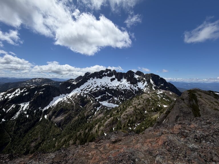 View from Mt. Cokely summit Vancouver Island BC showing snow-streaked mountain peaks and forested valley under blue sky